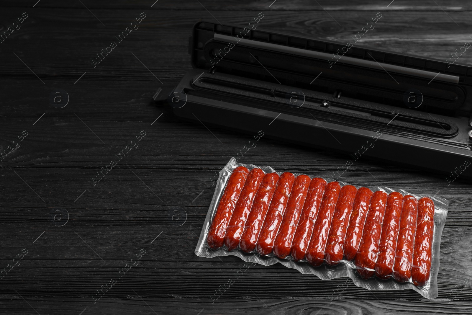 Photo of Vacuum packing sealer and plastic bag of sausages on black wooden table, space for text