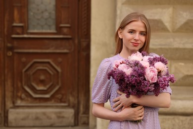 Beautiful woman with bouquet of spring flowers near building outdoors, space for text