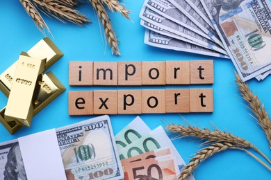 Photo of Words Import and Export made of wooden squares, ears of wheat, banknotes and gold bars on light blue background, flat lay