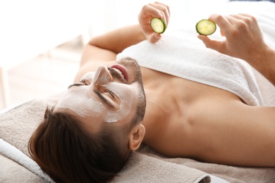 Photo of Young man with facial mask holding cucumber slices in spa salon