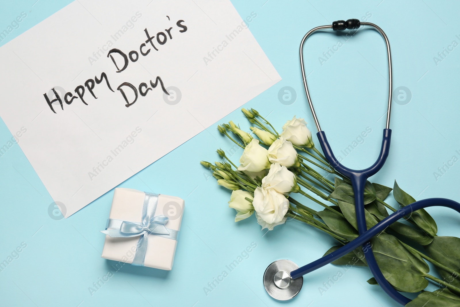 Photo of Card with phrase Happy Doctor's Day, stethoscope, gift box and eustoma flowers on light blue background, flat lay