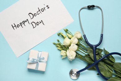 Photo of Card with phrase Happy Doctor's Day, stethoscope, gift box and eustoma flowers on light blue background, flat lay