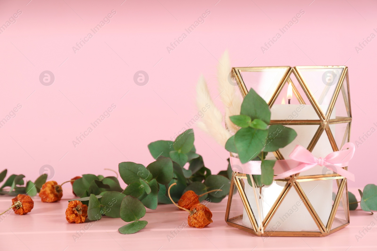 Photo of Stylish glass holder with burning candle and floral decor on pink wooden table. Space for text