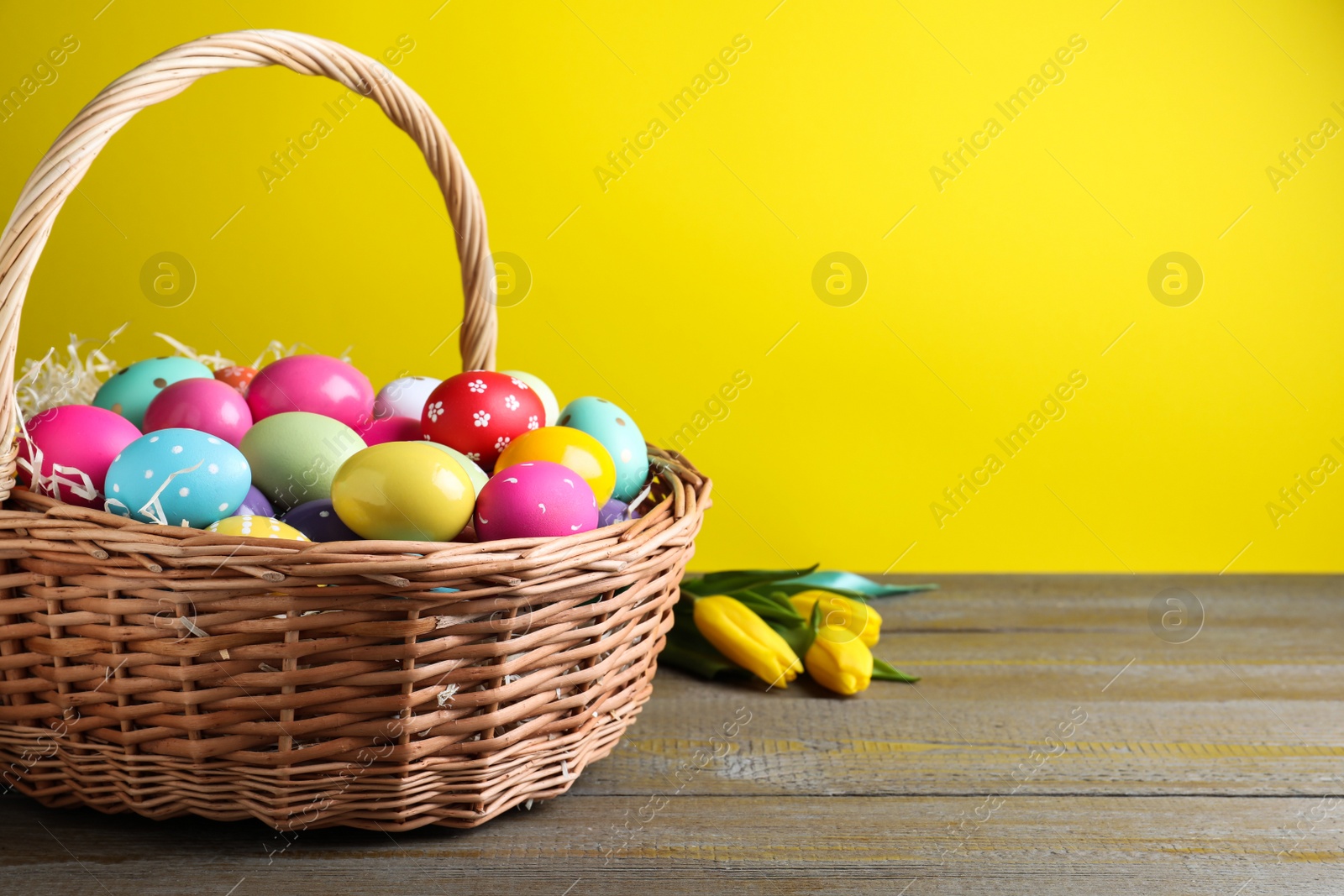 Photo of Colorful Easter eggs in wicker basket and tulips on wooden table against yellow background. Space for text