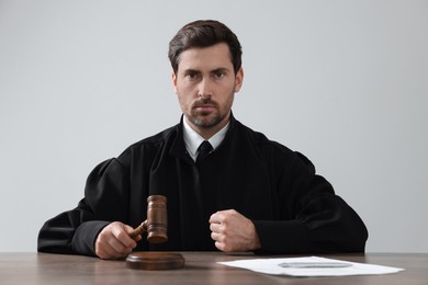 Judge with gavel and papers sitting at wooden table against light grey background
