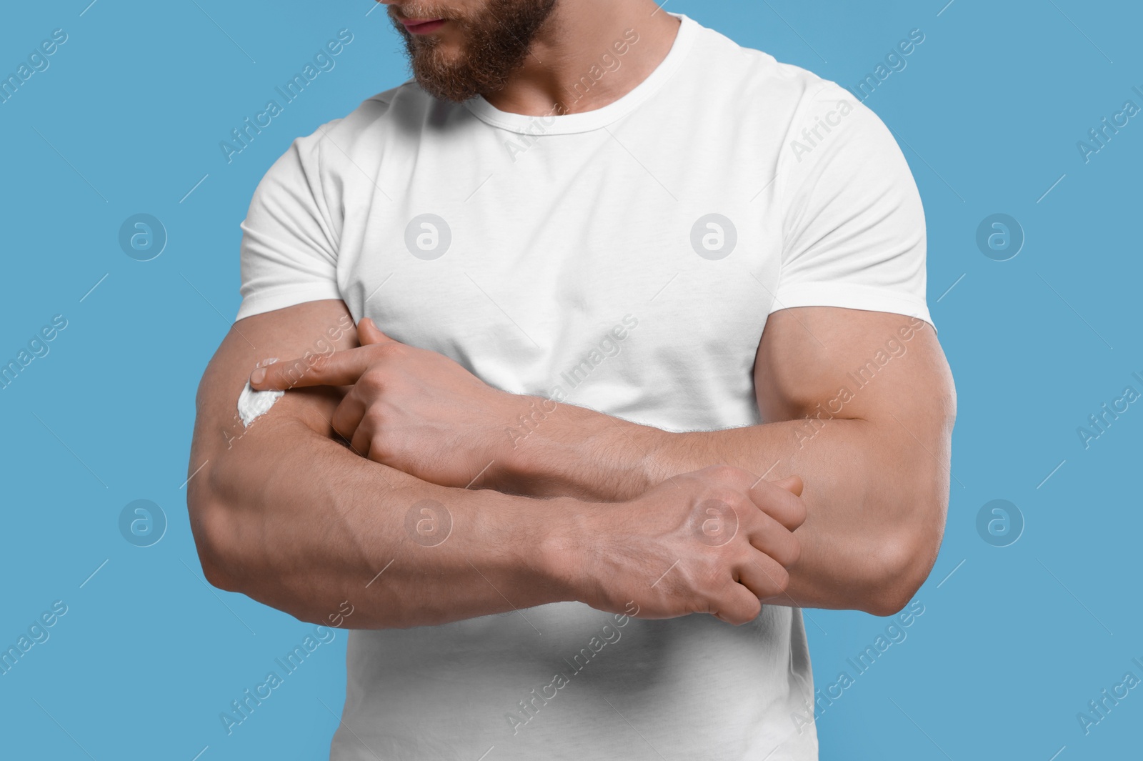 Photo of Man applying body cream onto his arm on light blue background, closeup