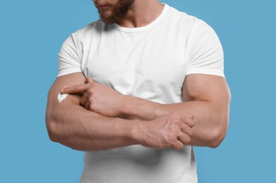 Man applying body cream onto his arm on light blue background, closeup