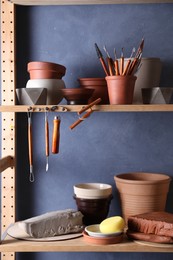 Photo of Set of different crafting tools and clay dishes on wooden rack in workshop