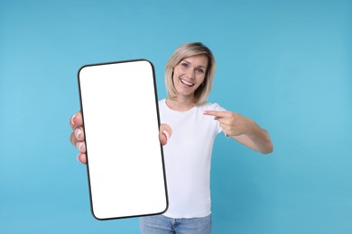 Happy woman pointing at mobile phone with blank screen on light blue background. Mockup for design
