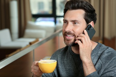 Handsome man with cup of coffee talking on phone indoors