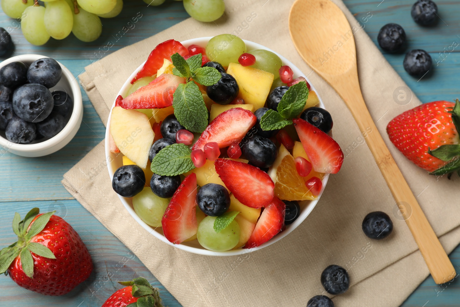 Photo of Tasty fruit salad in bowl, ingredients and spoon on light blue wooden table, flat lay