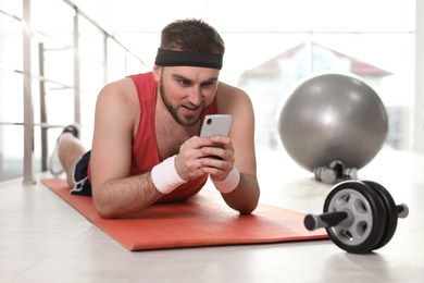 Photo of Lazy young man with sport equipment using smartphone lying on yoga mat