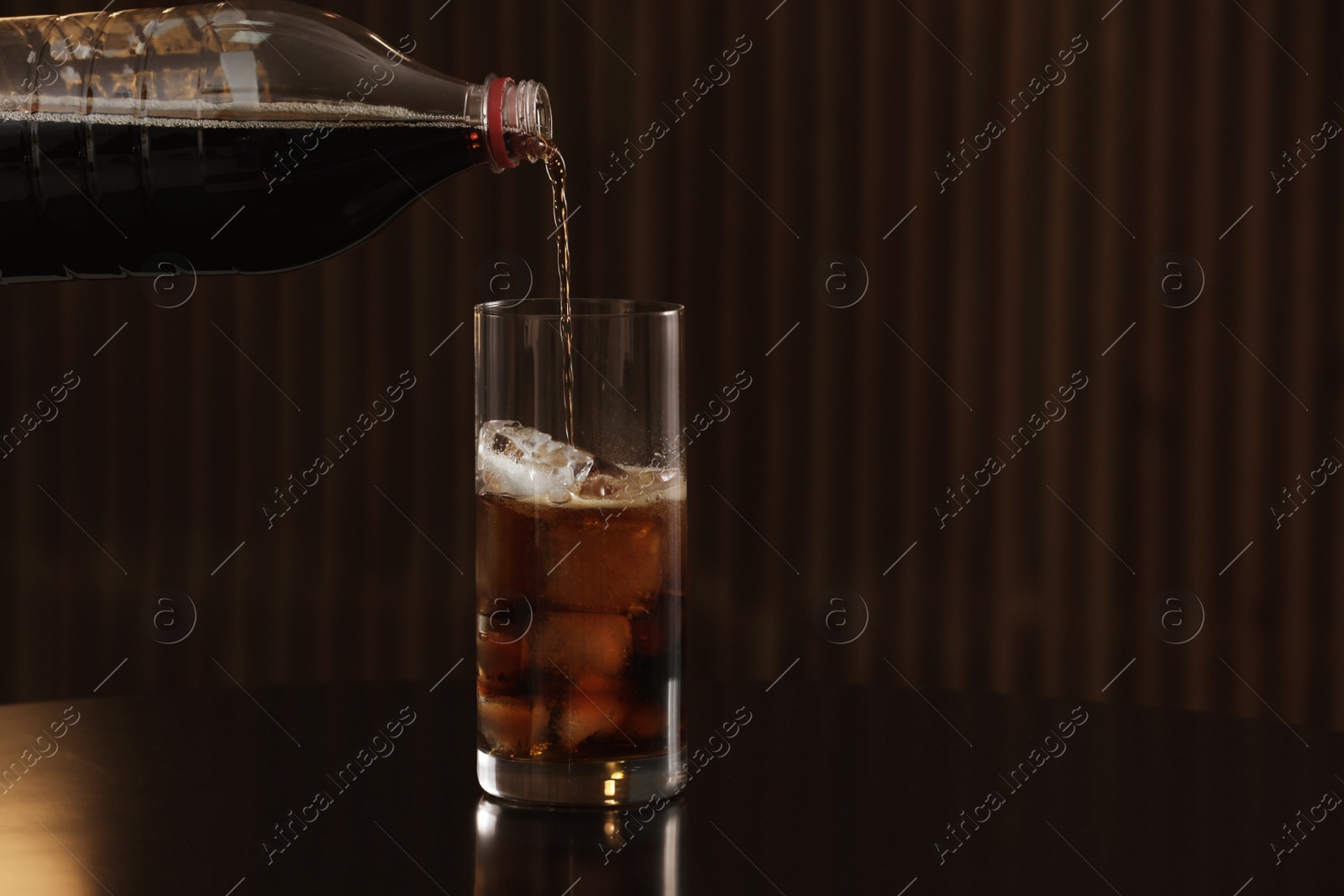 Photo of Pouring cola from bottle into glass with ice cubes on table against blurred background. Space for text