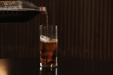 Pouring cola from bottle into glass with ice cubes on table against blurred background. Space for text