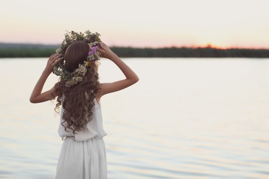 Photo of Little girl wearing wreath made of beautiful flowers near river at sunset