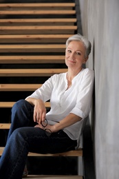 Photo of Happy mature woman sitting on wooden stairs indoors. Smart aging