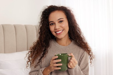 Happy African American woman with cup of drink in bedroom