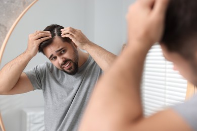Emotional man with dandruff in his dark hair near mirror indoors