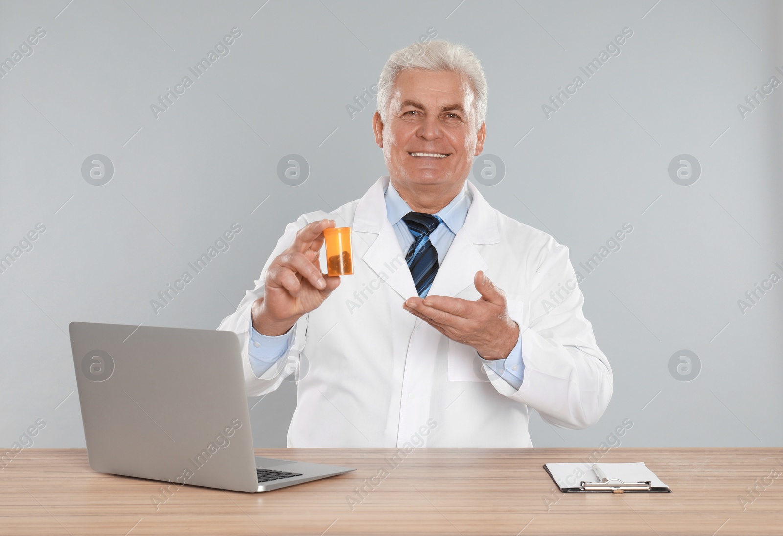 Photo of Professional pharmacist with pills and laptop at table against light grey background