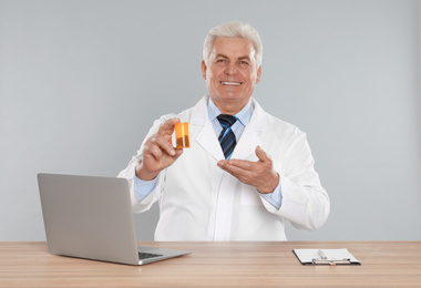 Photo of Professional pharmacist with pills and laptop at table against light grey background