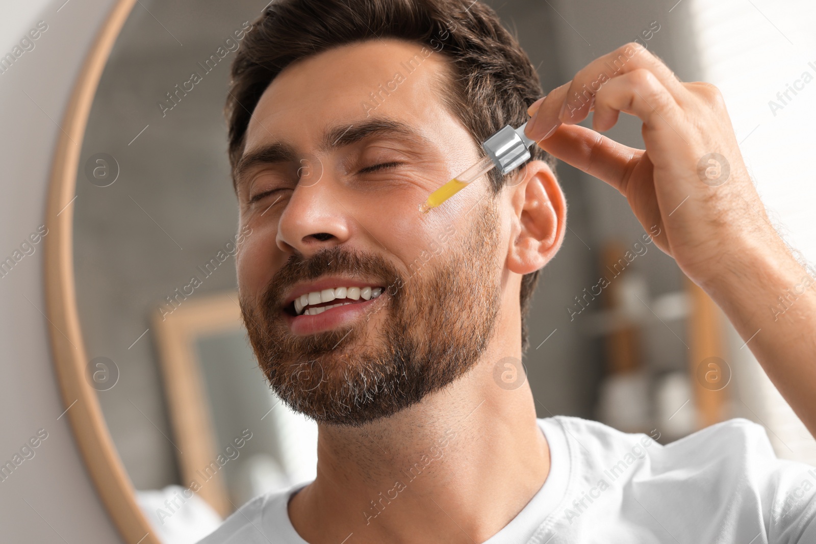 Photo of Smiling man applying cosmetic serum onto his face indoors. Space for text