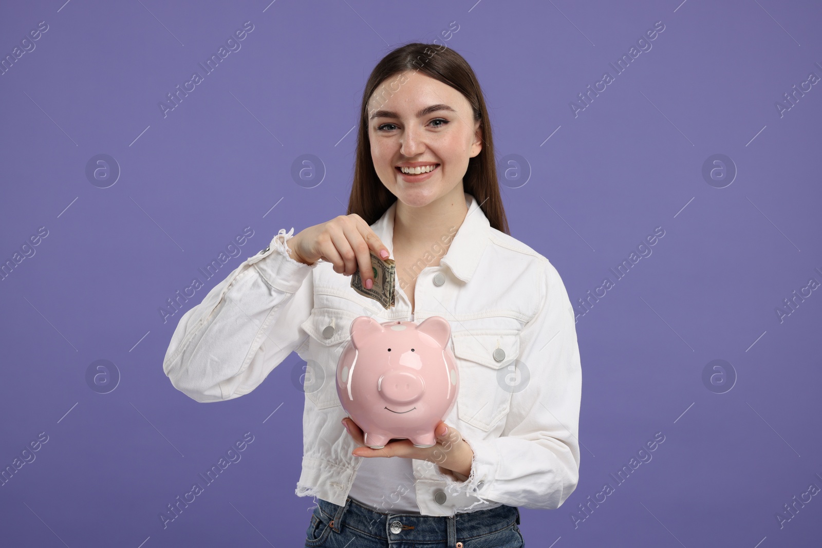 Photo of Happy woman putting dollar banknote into piggy bank on purple background