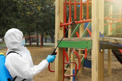 Woman wearing chemical protective suit with disinfectant sprayer on playground. Preventive measure during coronavirus pandemic