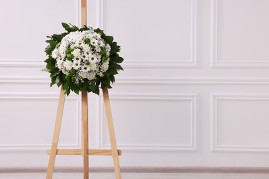 Funeral wreath of flowers on wooden stand near white wall indoors. Space for text