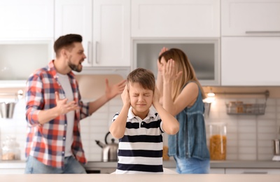 Little unhappy boy sitting at table while parents arguing on kitchen