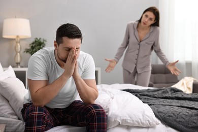 Photo of Stressed husband sitting on bed while his wife blaming him in bedroom, selective focus. Relationship problems