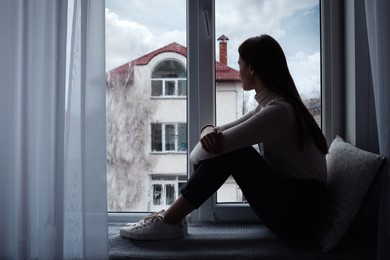 Photo of Melancholic young woman looking out of window indoors, space for text. Loneliness concept