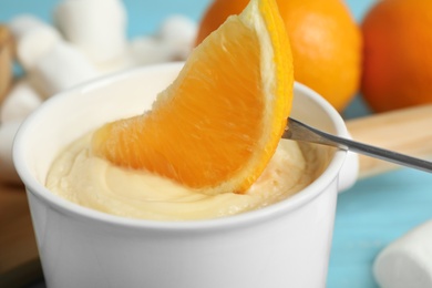 Dipping slice of orange into pot with white chocolate fondue, closeup