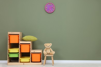 Shelves and teddy bear on chair near olive wall in playroom, space for text. Stylish kindergarten interior