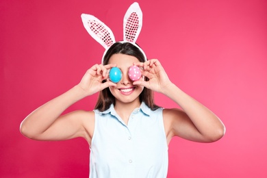 Beautiful woman in bunny ears headband holding Easter eggs near eyes on color background