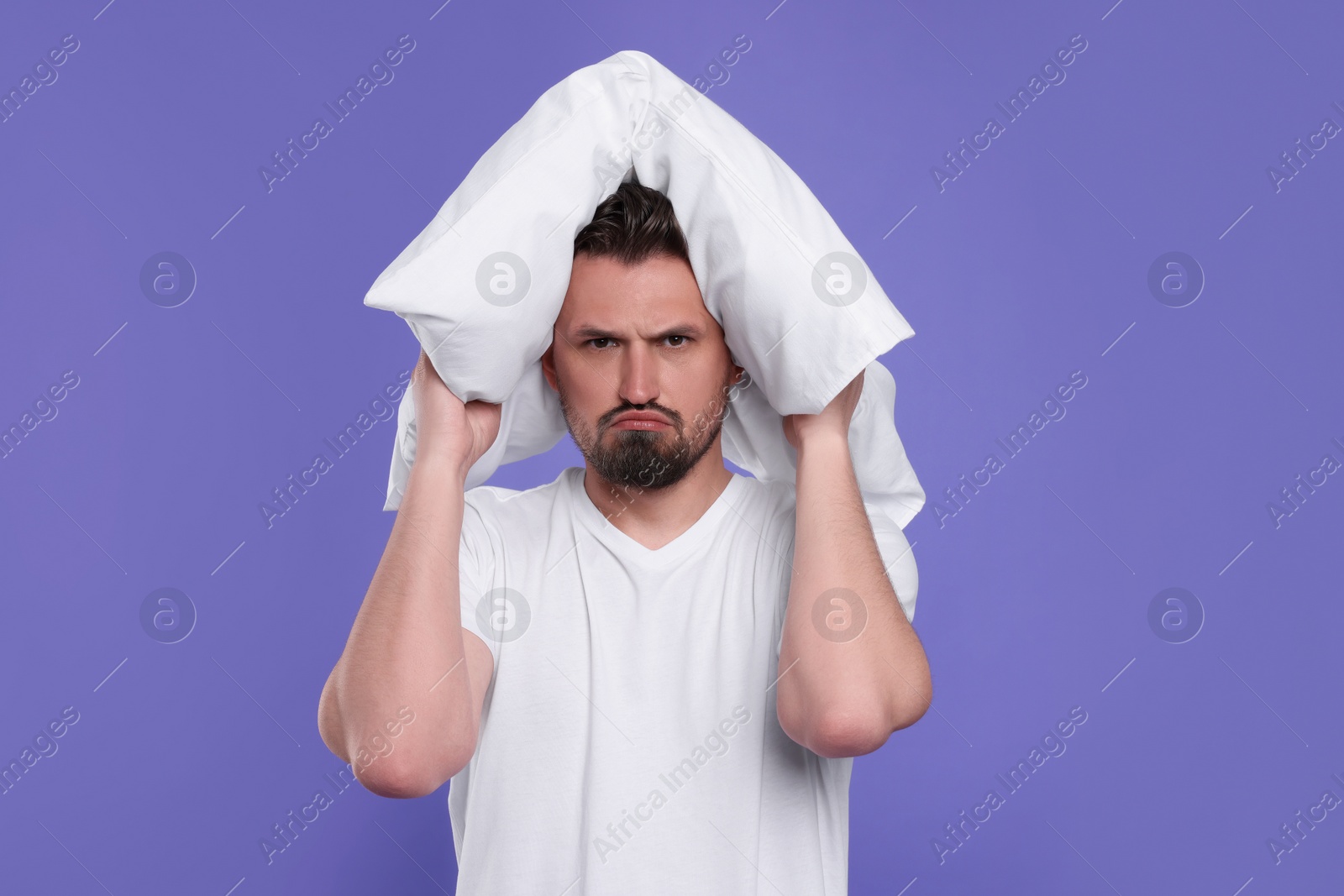 Photo of Unhappy man covering ears with pillow on violet background. Insomnia problem