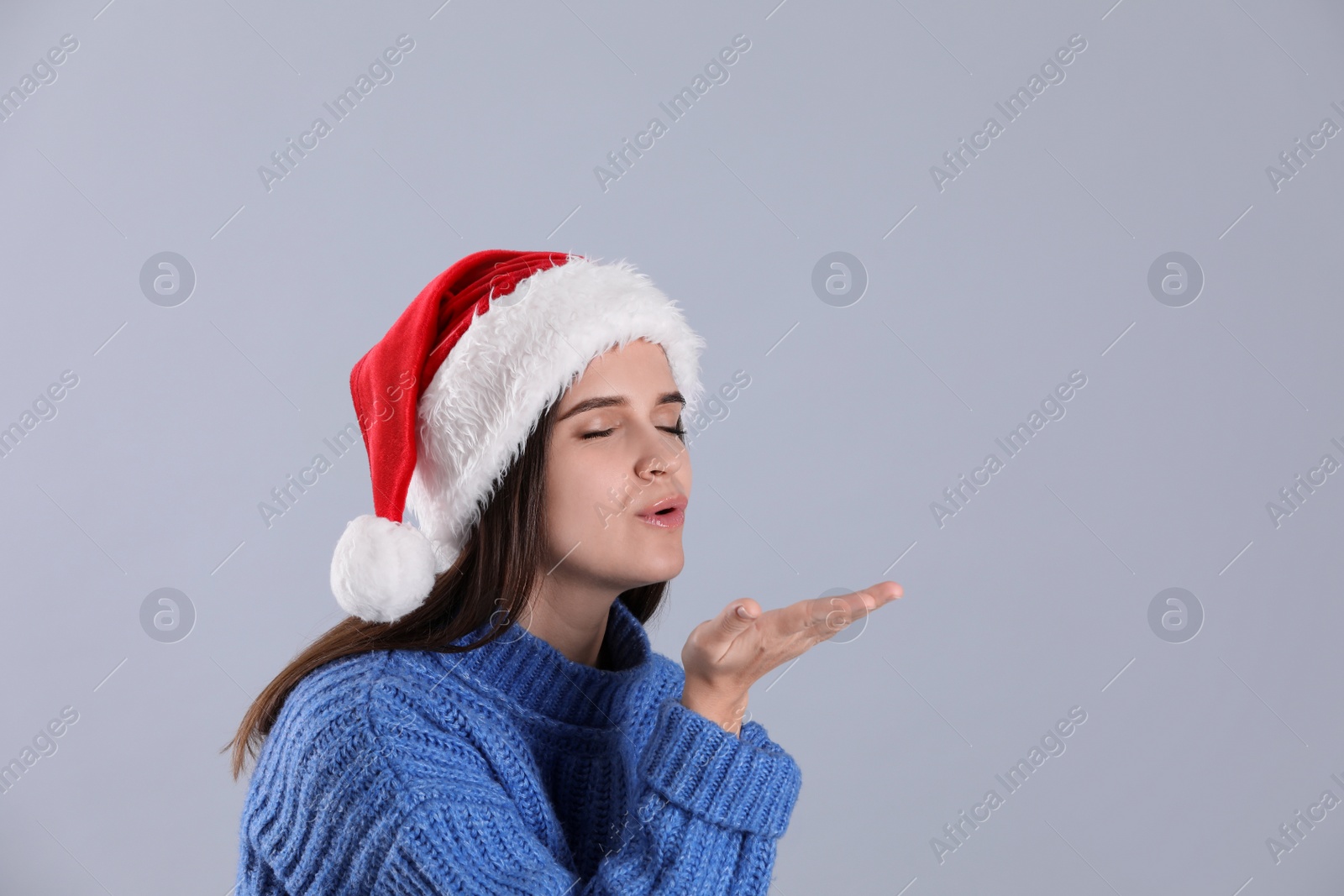 Photo of Pretty woman in Santa hat and blue sweater blowing kiss on grey background, space for text
