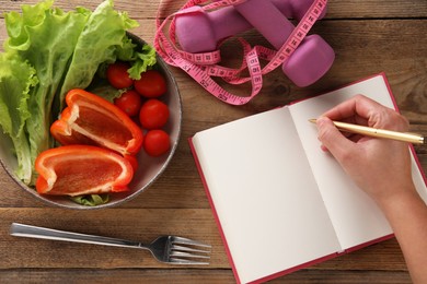 Photo of Woman developing diet plan at wooden table with products, measuring tape and dumbbells, top view