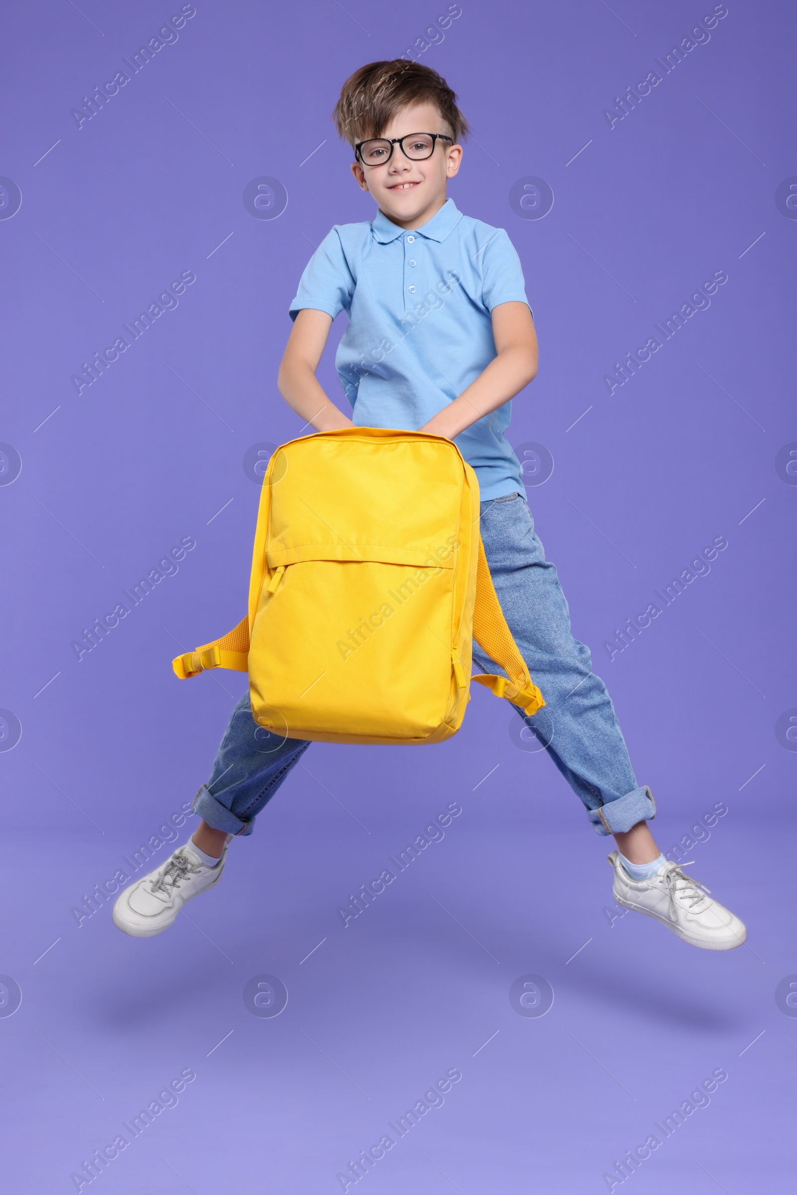 Photo of Cute schoolboy with backpack jumping on violet background, space for text