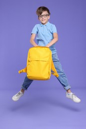 Cute schoolboy with backpack jumping on violet background, space for text