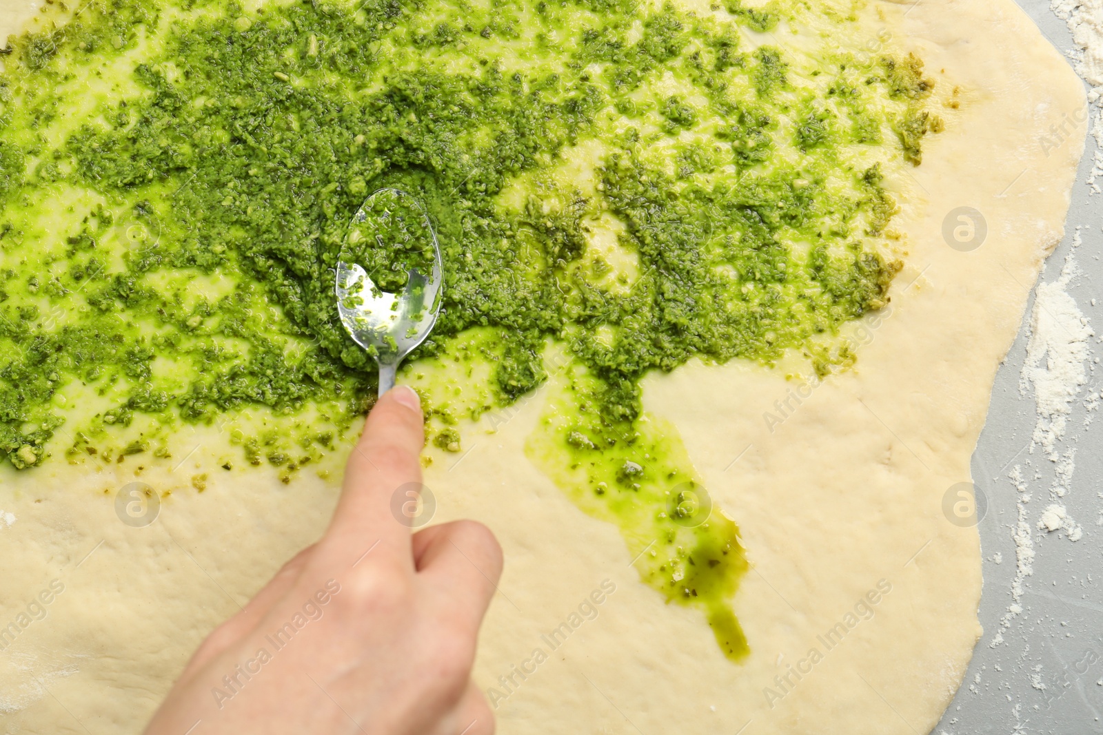Photo of Making delicious pesto bread. Woman putting sauce onto raw dough at grey table, top view