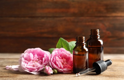 Photo of Bottles of rose essential oil, pipette and flowers on wooden table. Space for text