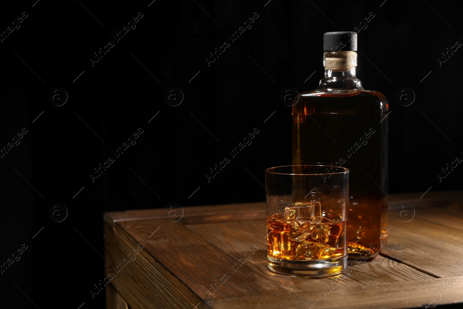 Photo of Whiskey with ice cubes in glass and bottle on wooden crate against black background, space for text