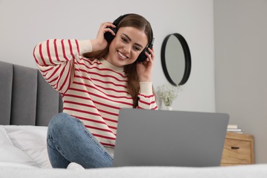 Photo of Happy woman with headphones and laptop on bed in bedroom