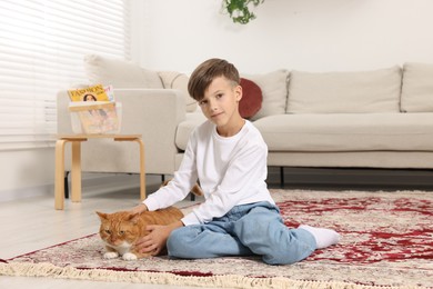 Photo of Little boy petting cute ginger cat on carpet at home