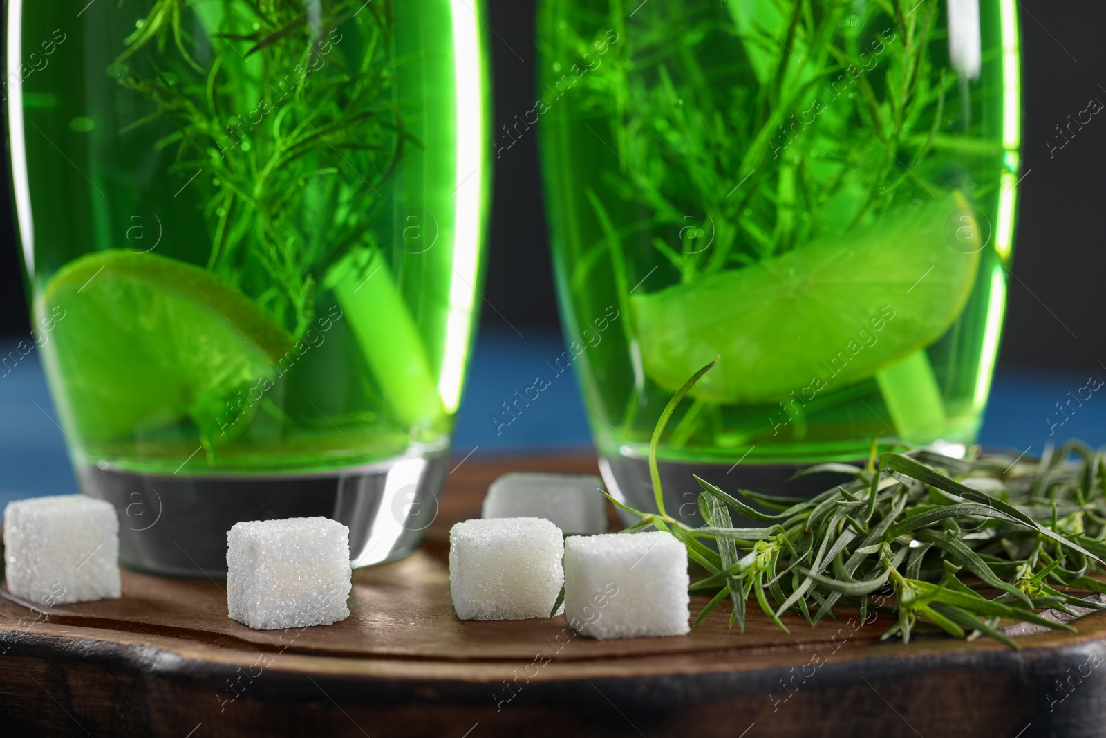 Photo of Glasses of homemade refreshing tarragon drink, sprigs and sugar cubes on wooden board, closeup