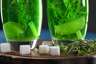 Glasses of homemade refreshing tarragon drink, sprigs and sugar cubes on wooden board, closeup