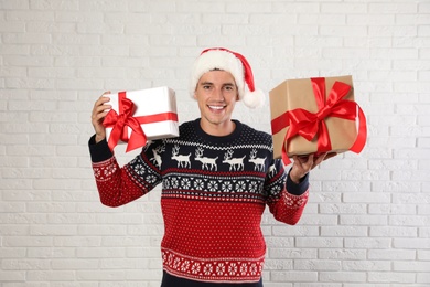 Happy man in Christmas sweater and Santa hat holding gift boxes near white brick wall