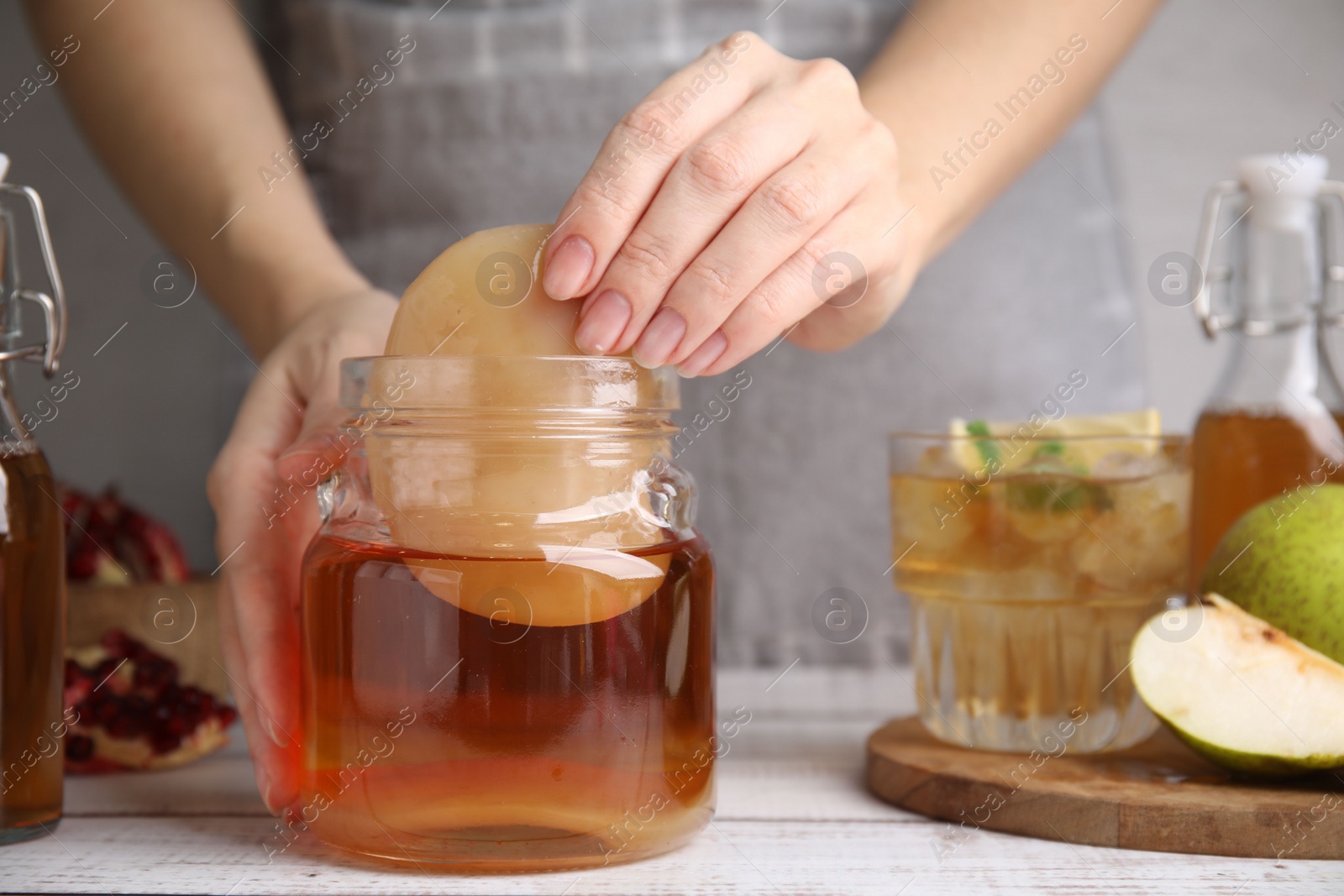 Photo of Woman putting Scoby fungus into jar with kombucha at white wooden table, closeup