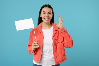 Photo of Happy young woman with blank white flag showing OK gesture on light blue background. Mockup for design