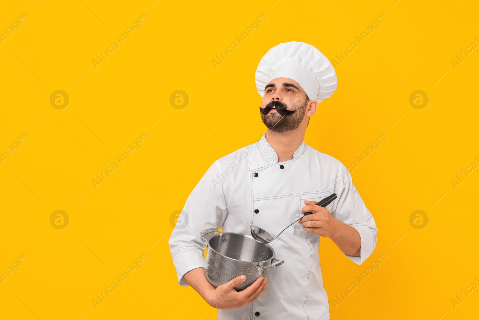 Photo of Professional chef with funny artificial moustache holding cooking pot and ladle on yellow background. Space for text
